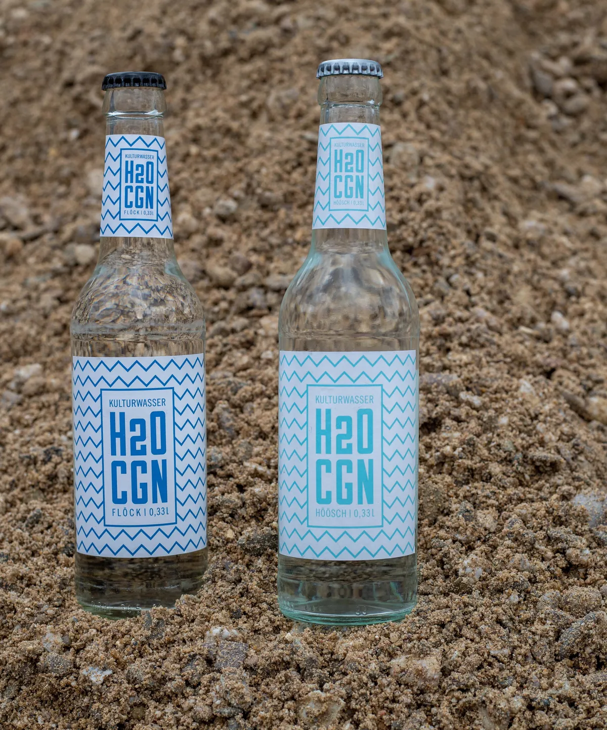 Zwei Flaschen H2O CGN, Flöck und Höösch, stehen im Sand